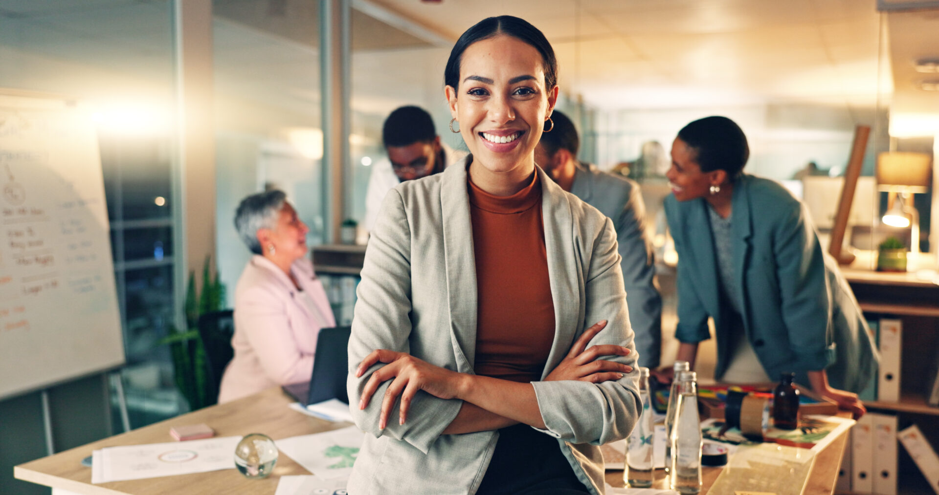 Confident businesswoman smiling with arms crossed in a busy office.