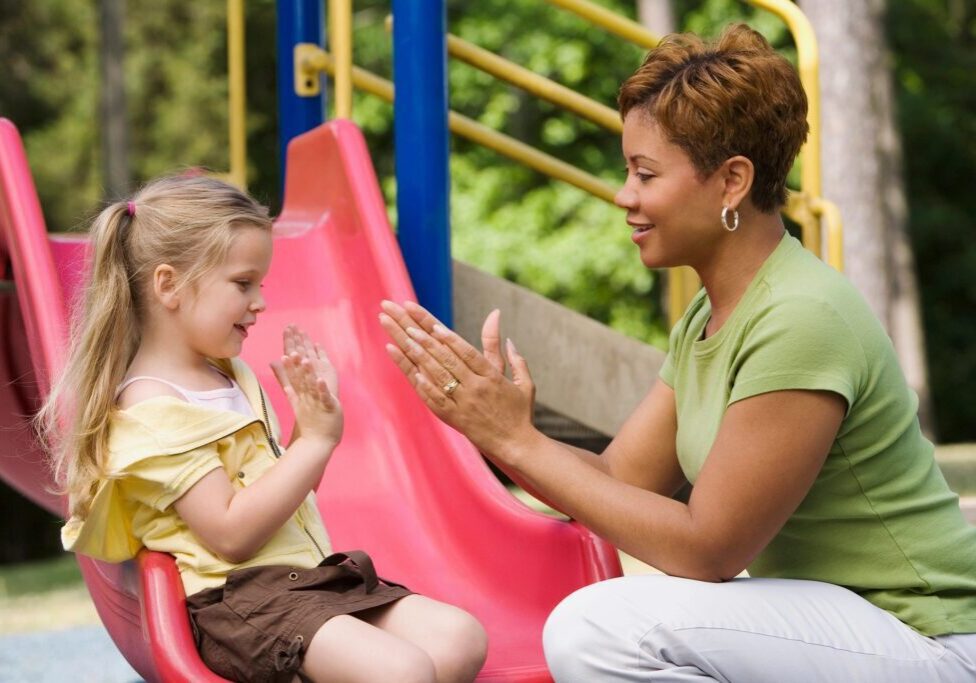 A woman and young girl happily playing clapping hands at a playground slide.